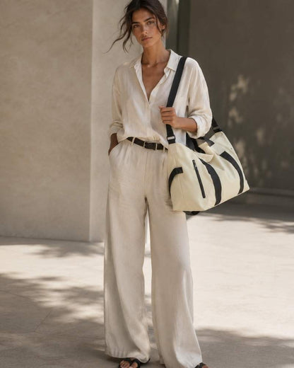 Model trägt ein weißes Leinenhemd und eine weite Leinenhose mit beige-schwarzer Damen Reisetasche in moderner Sommerkulisse.
