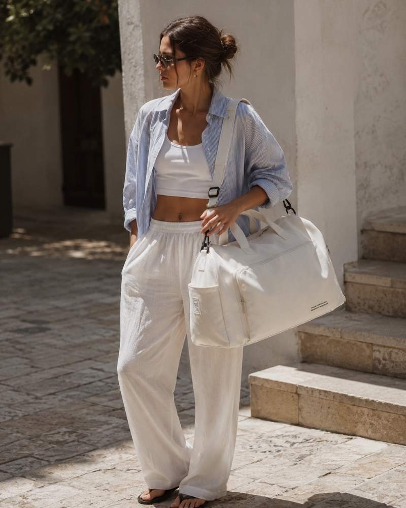 Model mit weißer Damen Reisetasche, lockerem blauem Hemd, weißem Crop-Top und weiter Leinenhose in mediterraner Kulisse.
