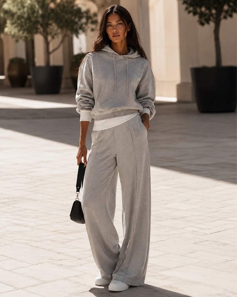 Model trägt einen grauen Damen Hoodie mit passender weiter Jogginghose und weißen Sneakern in moderner urbaner Kulisse.
