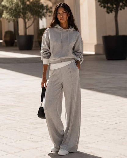 Model trägt einen grauen Damen Hoodie mit passender weiter Jogginghose und weißen Sneakern in moderner urbaner Kulisse.
