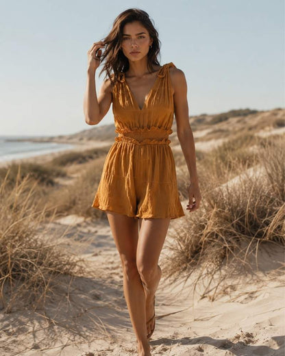 Model trägt einen ockerfarbenen ärmellosen Damen Jumpsuit mit V-Ausschnitt, Schleifenträgern und Shorts in den Sanddünen am Meer.
