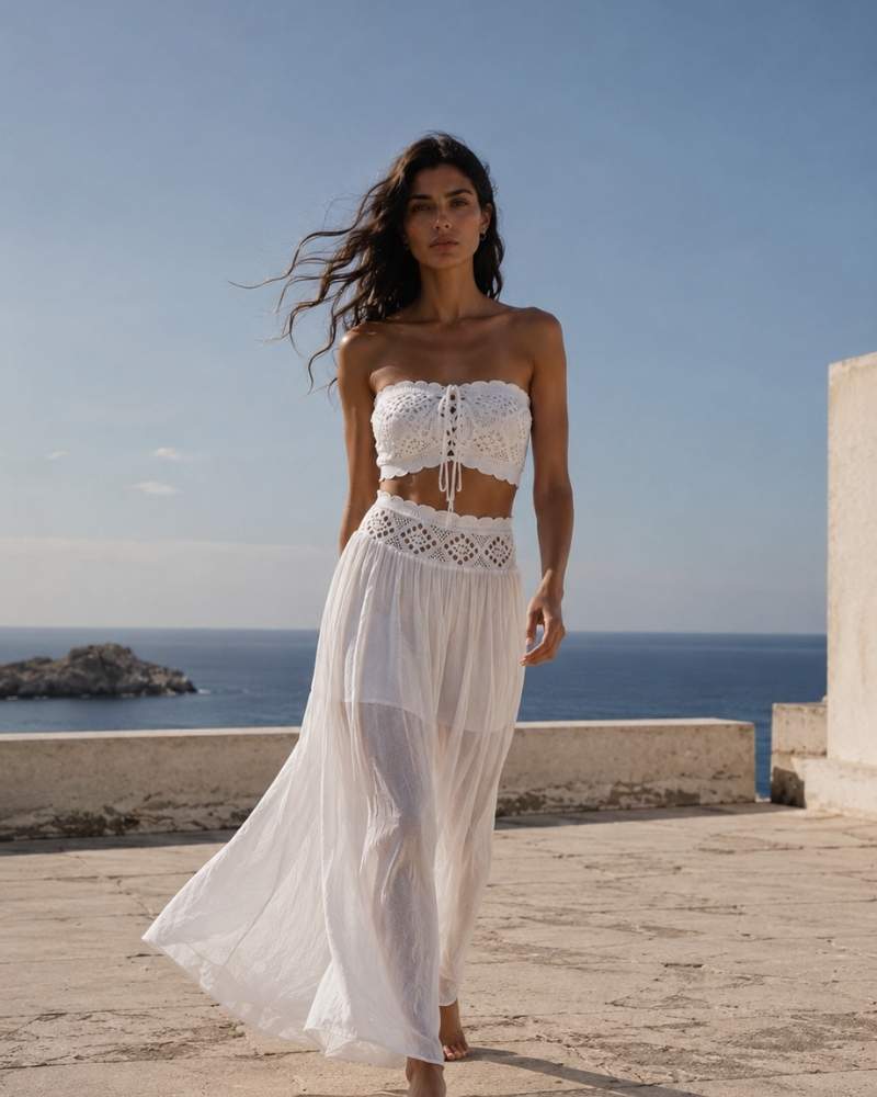 Model trägt ein weißes gehäkeltes Bandeau-Top und einen transparenten weißen Maxirock auf einer sonnigen Terrasse mit Meerblick.

