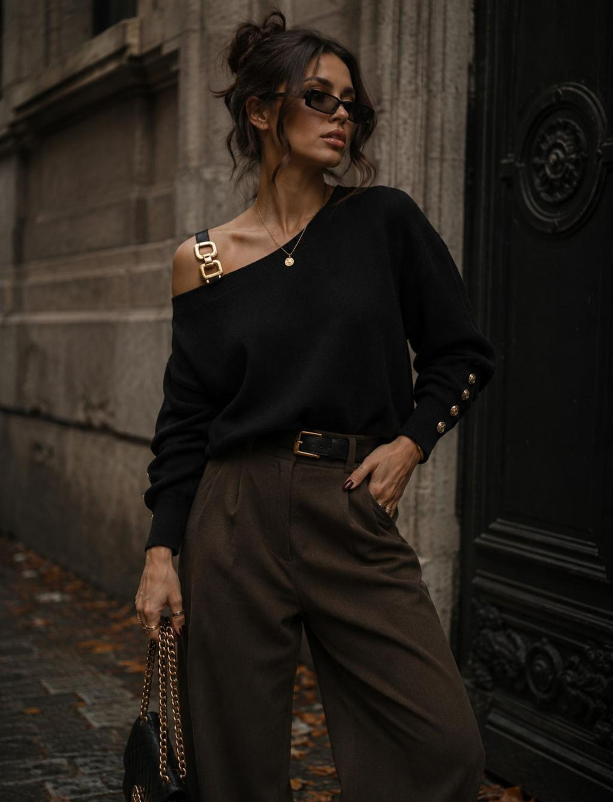 Damen Rebel Herbst Outfit mit schwarzem One Shoulder Pullover mit goldenen Details, brauner weiter Hose und schwarzer Handtasche