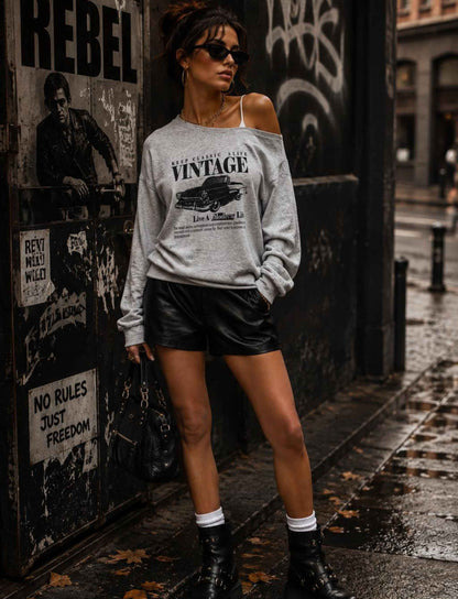 Grauer Vintage-Pullover für Damen im Off-Shoulder-Look kombiniert mit schwarzen Leder-Shorts und Boots im herbstlichen Rebel Streetstyle